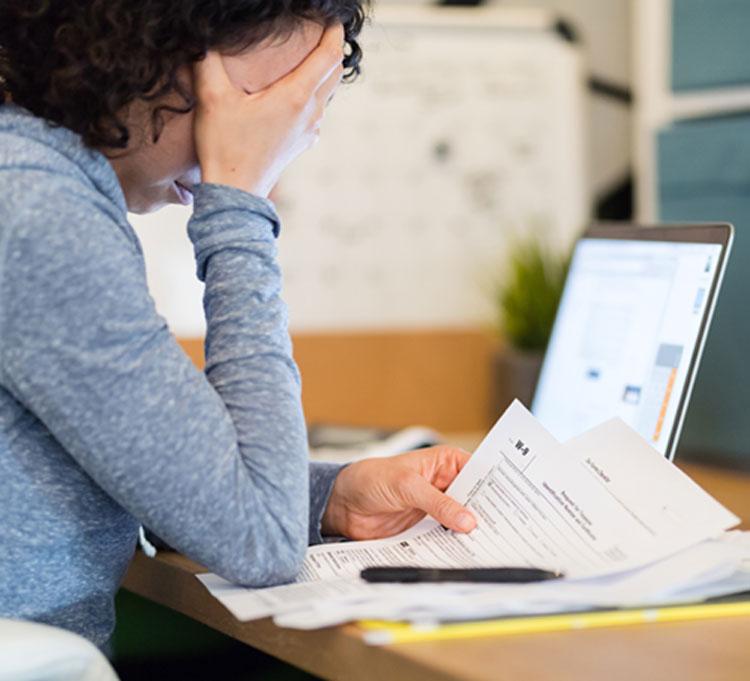 Stressed woman wearing a blue sweater, sitting at a desk reviewing unpaid taxes, with her computer open
