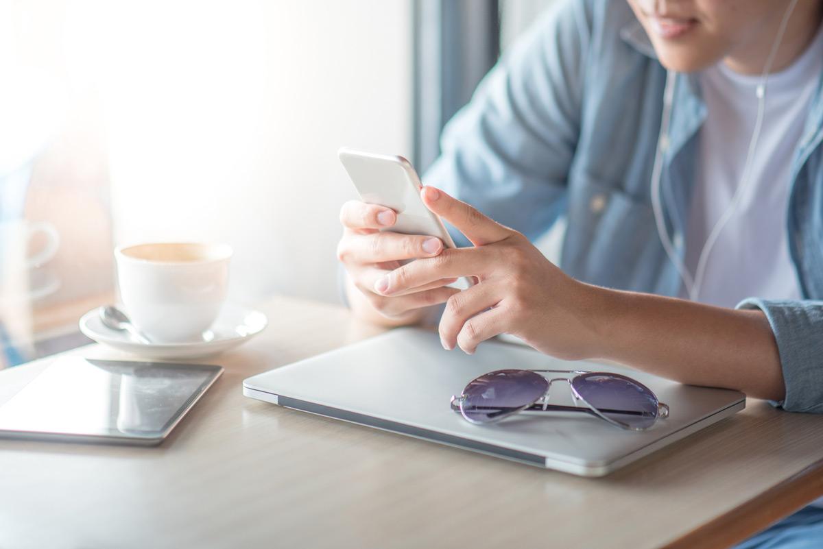 Photo of a man sitting at a desk with a laptop, phone, and a cup of coffee, using phone to message